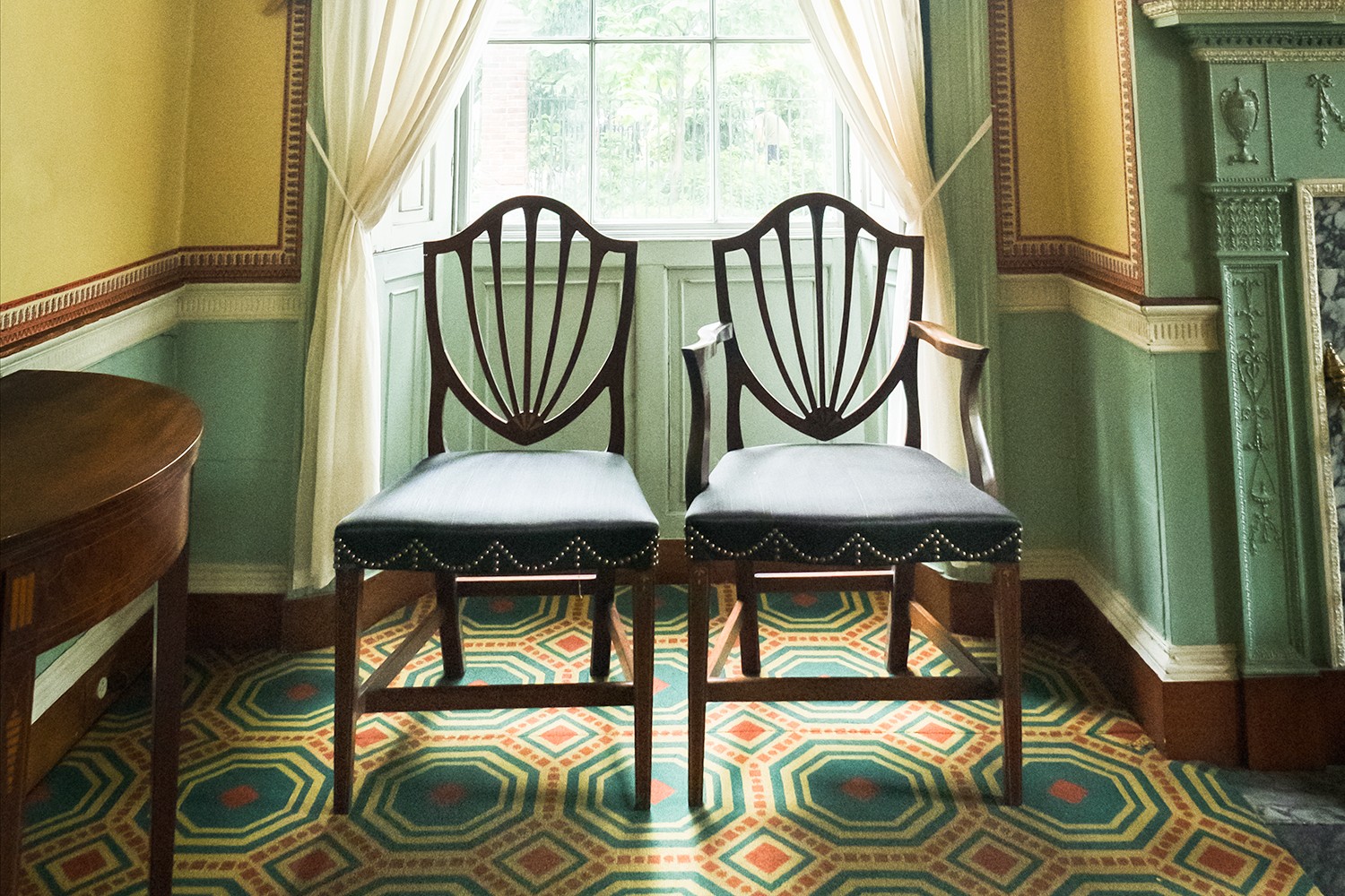 Two dark-wood chairs with a shield-shaped back, the one on the right has arms, placed in front of a window and framed by sheer curtains. The surrounding walls are a grey-green painted color on the bottom with a yellow wallpaper with a red border at the to